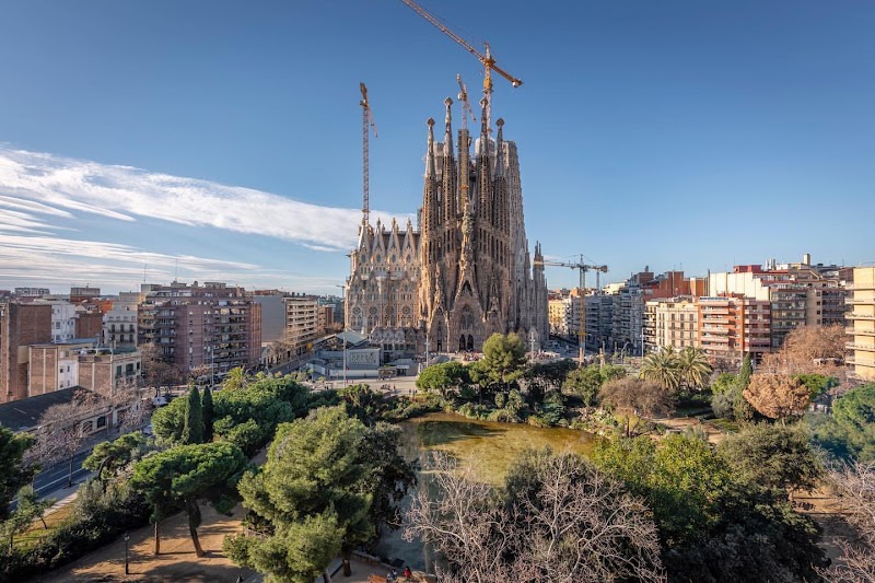 Aerial view of La Sagrada Família in Barcelona, seamlessly blending into the city's urban fabric and greenery, with cranes rising above the basilica. This iconic landmark is a must-see, prominently featured on any comprehensive tourist map of the area.