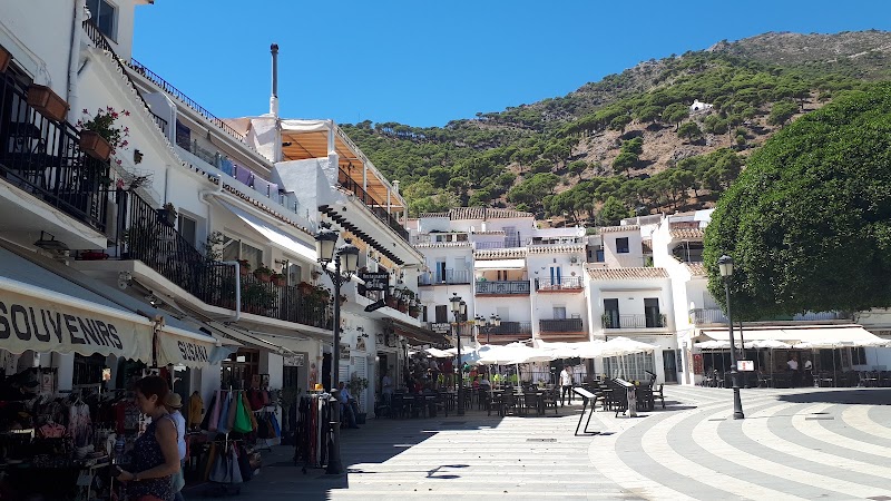 A street lined with souvenir shops and outdoor seating, where white buildings stand against green hills under a clear blue sky, offers a charming spot marked on every tourist map.