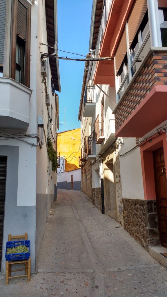 Narrow alleyway between residential buildings with balconies on a sunny day. An orange building is visible at the end of the alley, a quaint spot often highlighted in travel maps. A small plant is near the entrance.