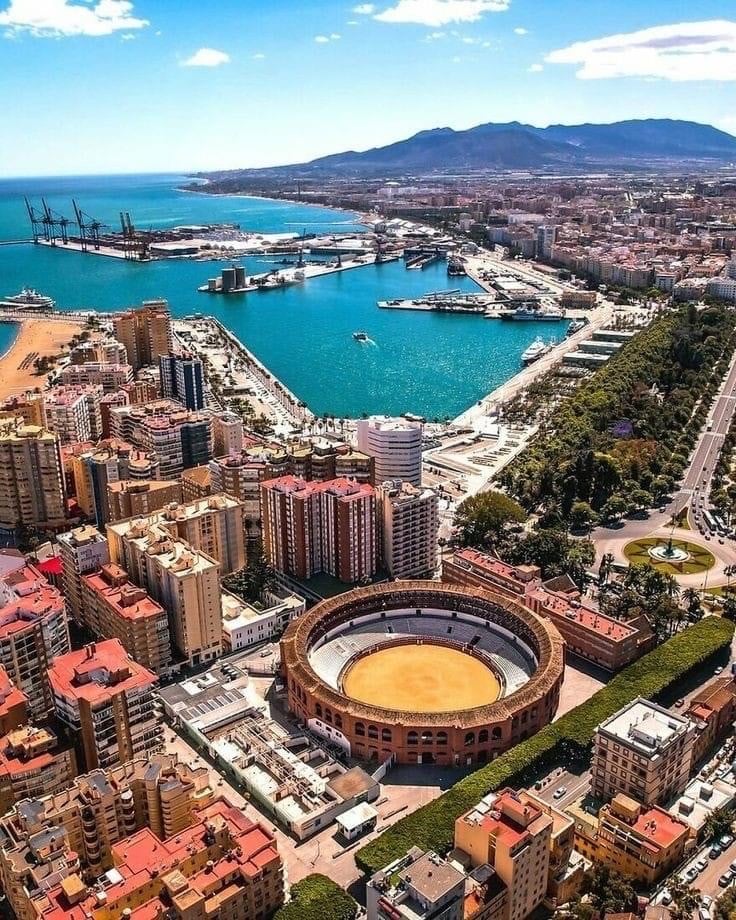 Aerial view of a coastal city featuring a circular bullring in the foreground, a bustling port with docks and cranes, and hills in the background under a blue sky, perfect for any travel map highlighting key attractions and scenic vistas.