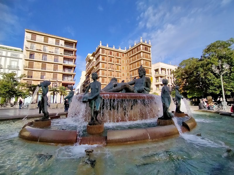 Fountain with statues in a city plaza, surrounded by buildings and trees under a blue sky—a perfect spot marked on any tourist map for those exploring the area.