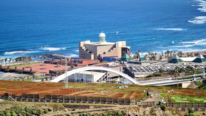 View of a modern architectural complex by the ocean, featuring a unique building with a white dome, surrounded by tennis courts and a prominent curved white bridge in the foreground—a perfect spot to mark on your travel map for an unforgettable experience.