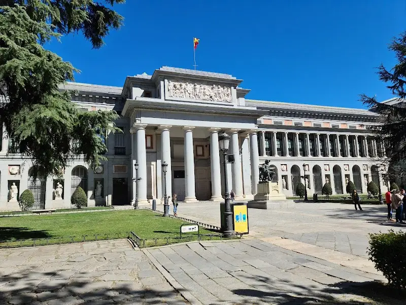 Front view of a neoclassical building with large columns, ornate carvings, and a flag on top. A few people clutching their tourist maps wander the plaza, while trees frame the scene under a clear blue sky.