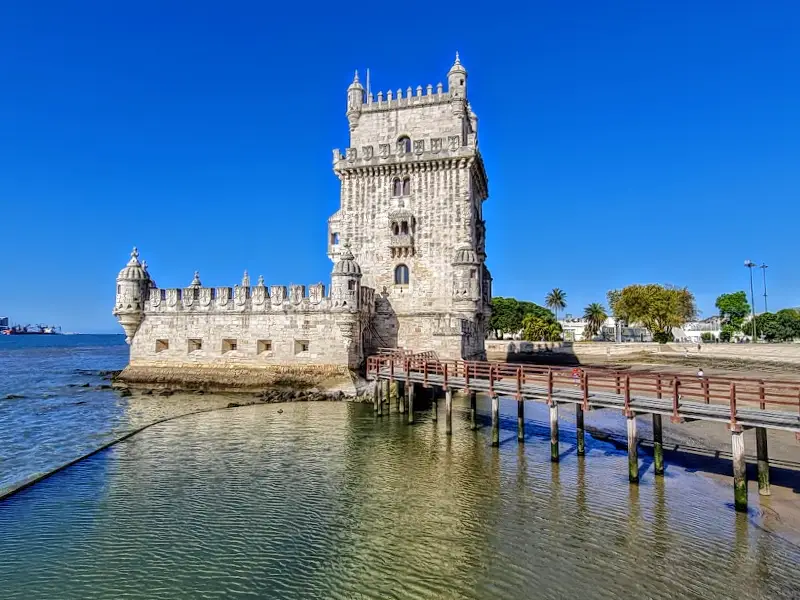 Historic stone tower with battlements by the water, connected to land by a wooden bridge, under a clear blue sky—perfectly framed on your travel map.