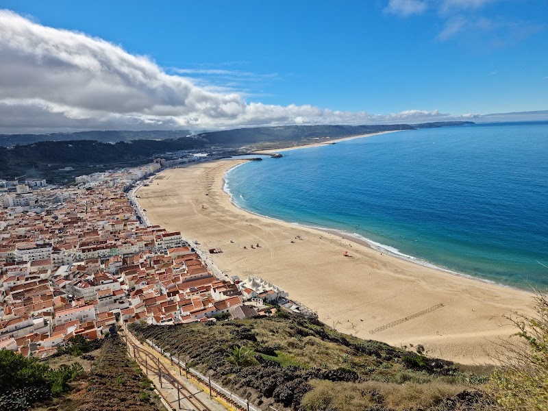 Aerial view of a coastal town with red-roofed buildings and a wide sandy beach curving alongside the blue ocean under a partly cloudy sky, resembling a vibrant tourist map.