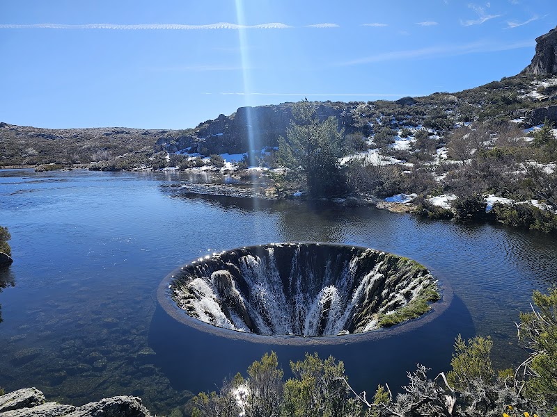 A funnel-shaped water sinkhole in a lake surrounded by rocky terrain and vegetation under a clear blue sky, inviting exploration with the help of a travel map.