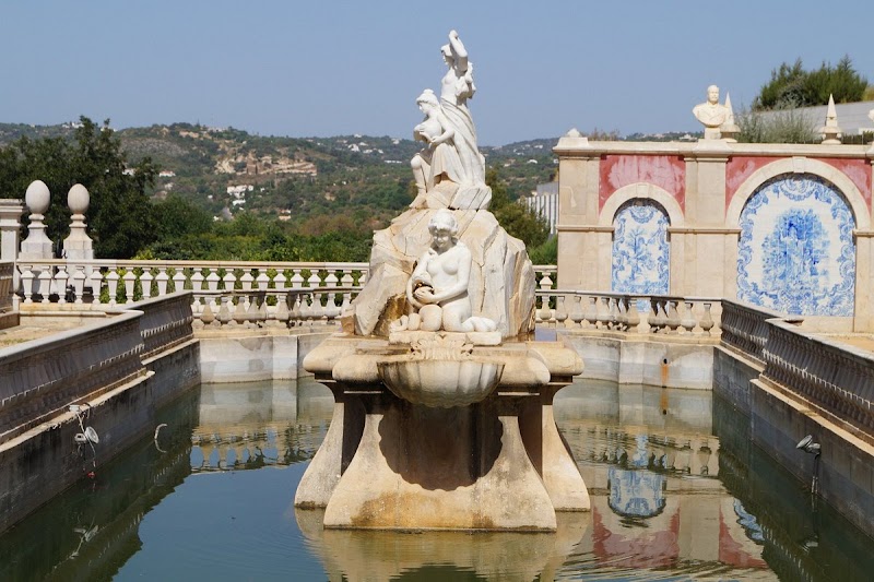 A stone fountain adorned with classical sculptures graces a reflecting pool, enclosed by a balustrade. Hills and trees form the backdrop under a clear sky, offering the perfect scene from a travel map.